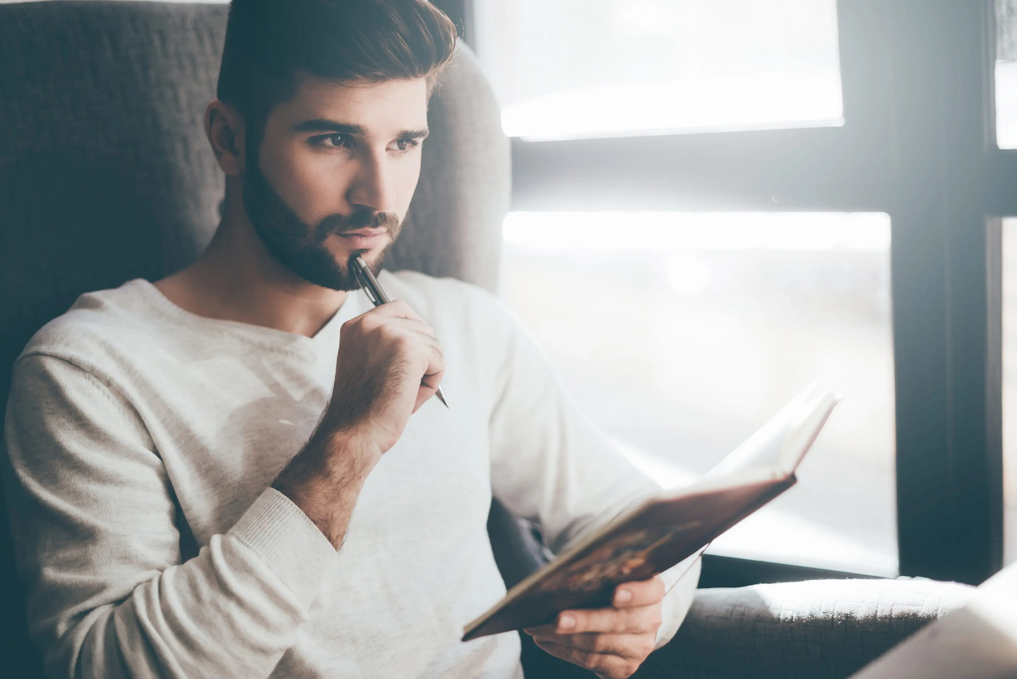 Man sitting in a chair, holding a pen in one hand and a book in the other