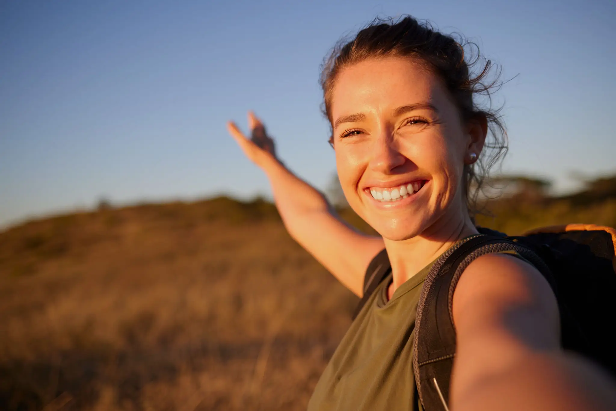 Smiling woman hiker takes a sunny selfie in a golden field, backpack visible, one arm outstretched; warm light conveys joy and adventure.