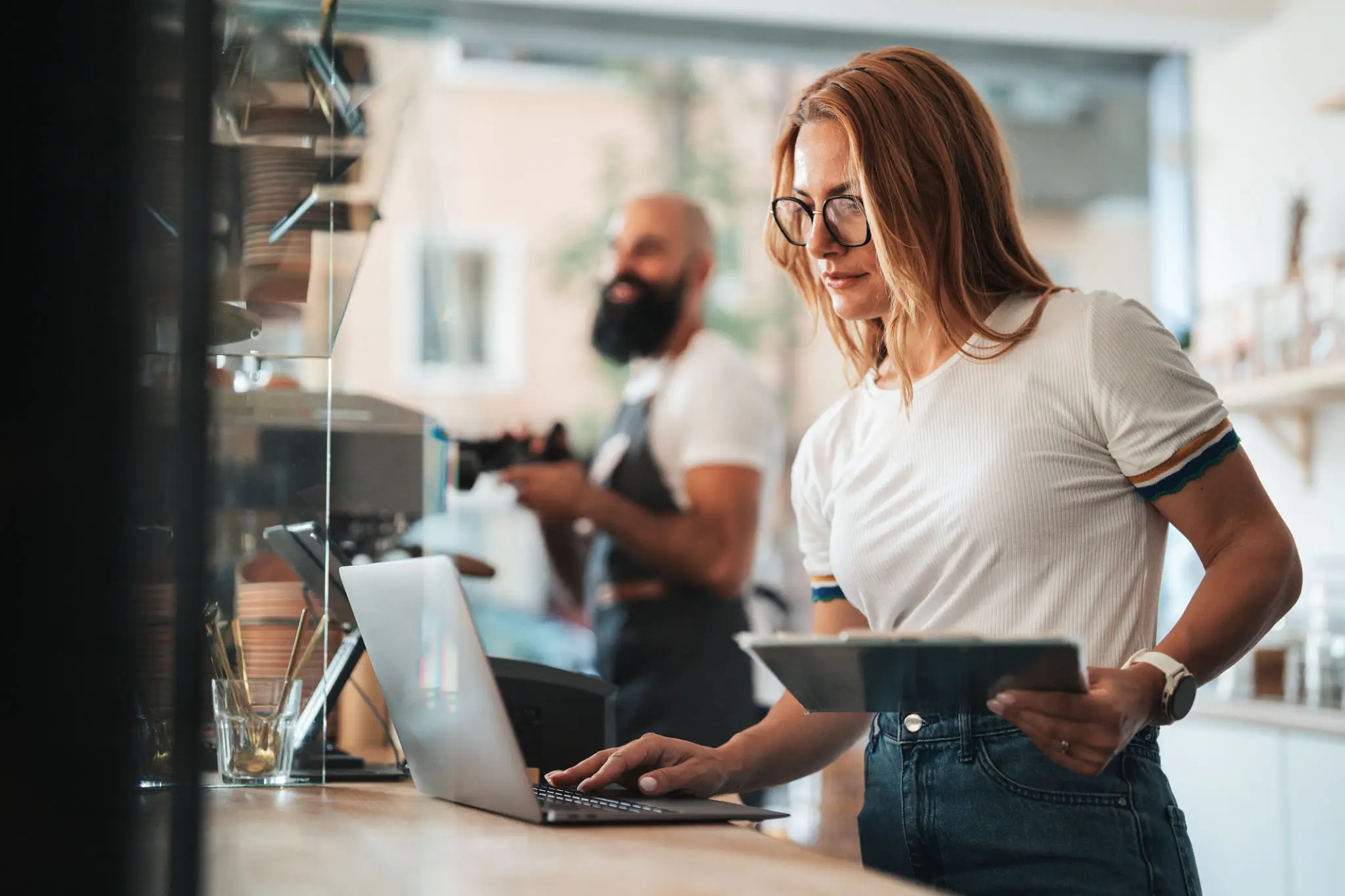Focused café worker with glasses uses a laptop and tablet behind the counter, while a bearded colleague works in the softly lit background.