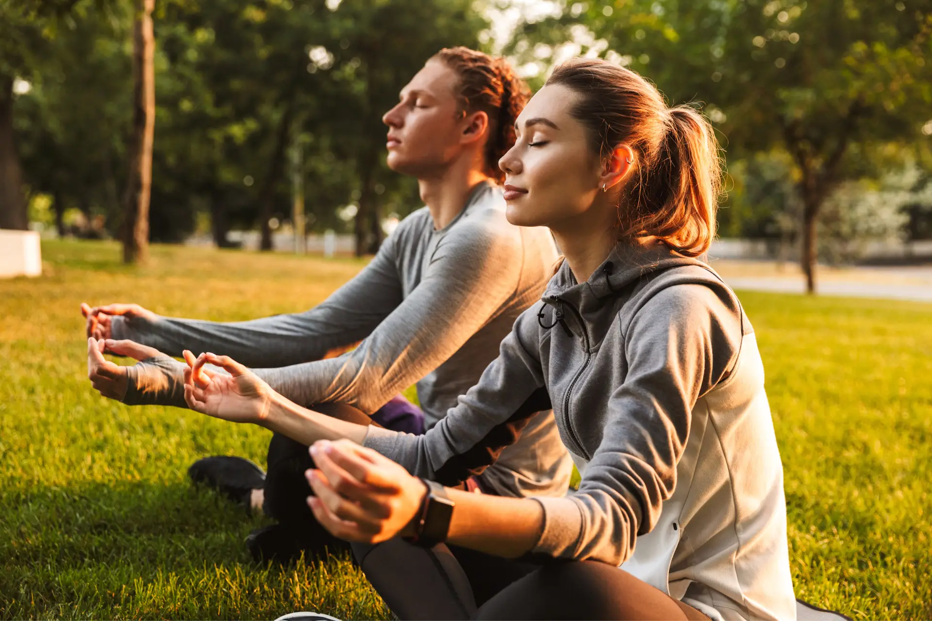 Man and woman meditating in the park, surrounded by lush green grass and tall trees