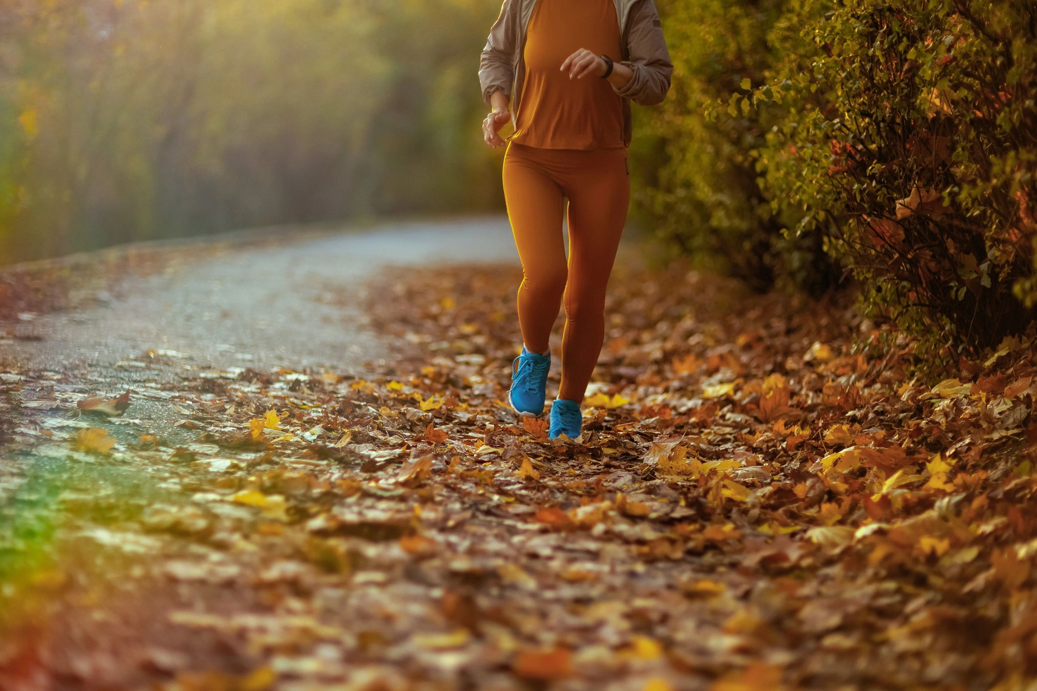 Woman jogging down a path in the woods, surrounded by trees and leaves on the ground