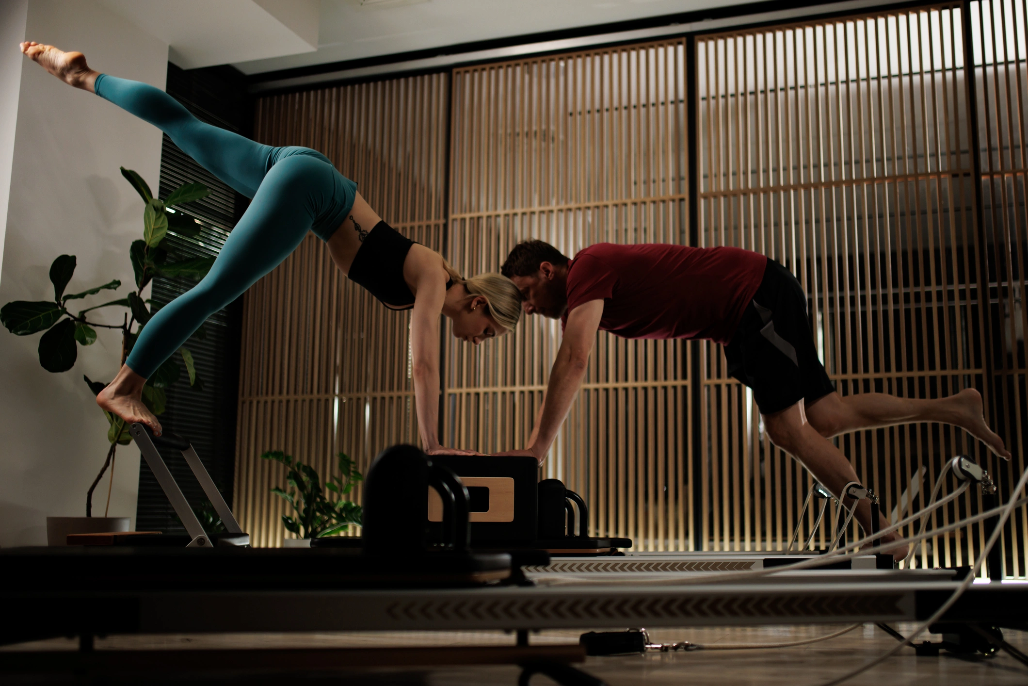 Man and woman doing pilates on a reformer in a studio, surrounded by plants and a wall