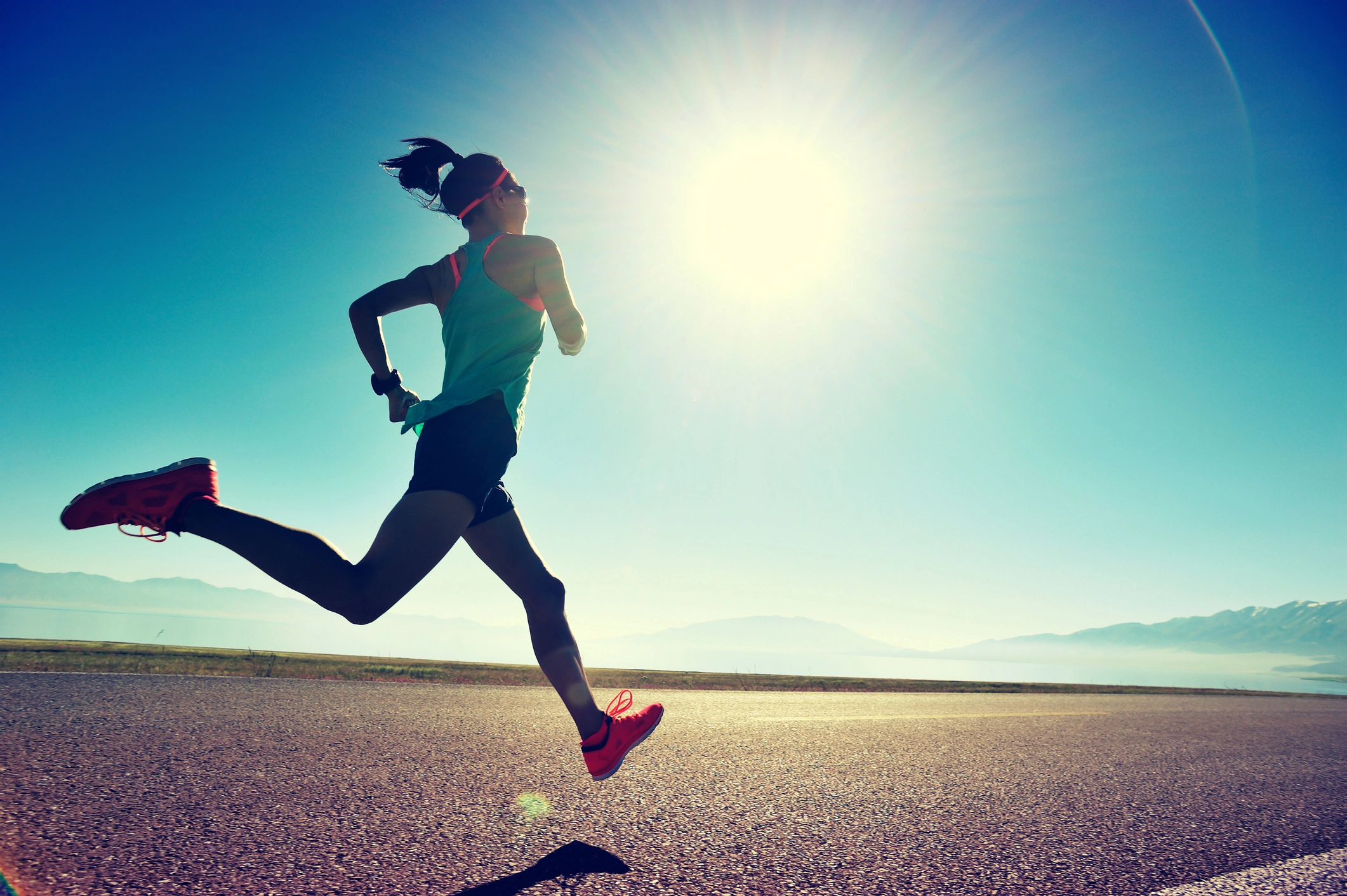 Woman running on a road with the sun shining brightly in the background, surrounded by lush green grass