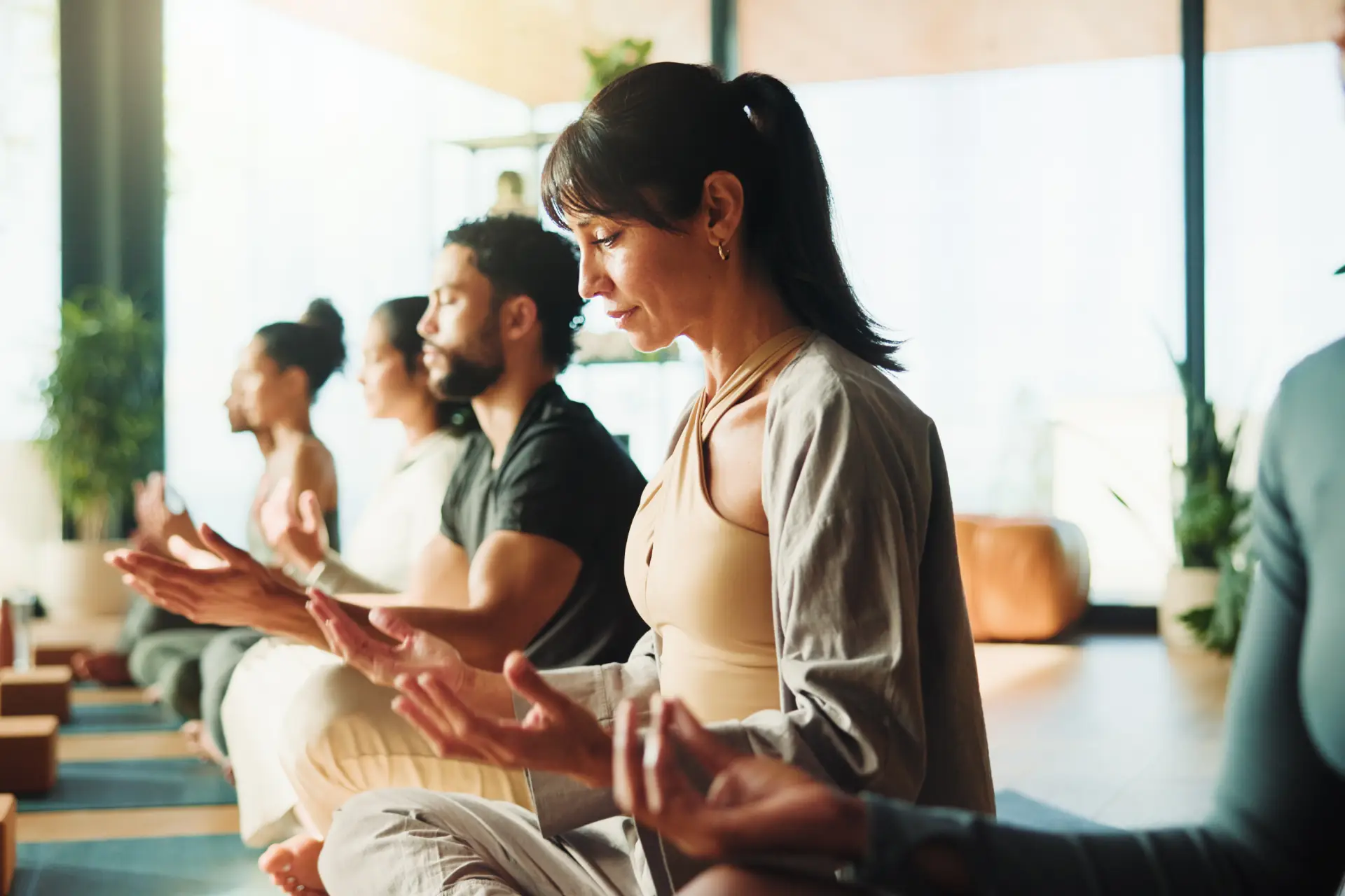 Group of people sitting in a yoga class, meditating on the floor surrounded by plants