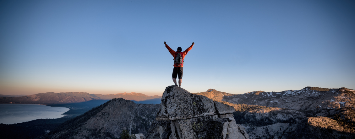Man standing atop a mountain with his arms outstretched, wearing a jacket and pants