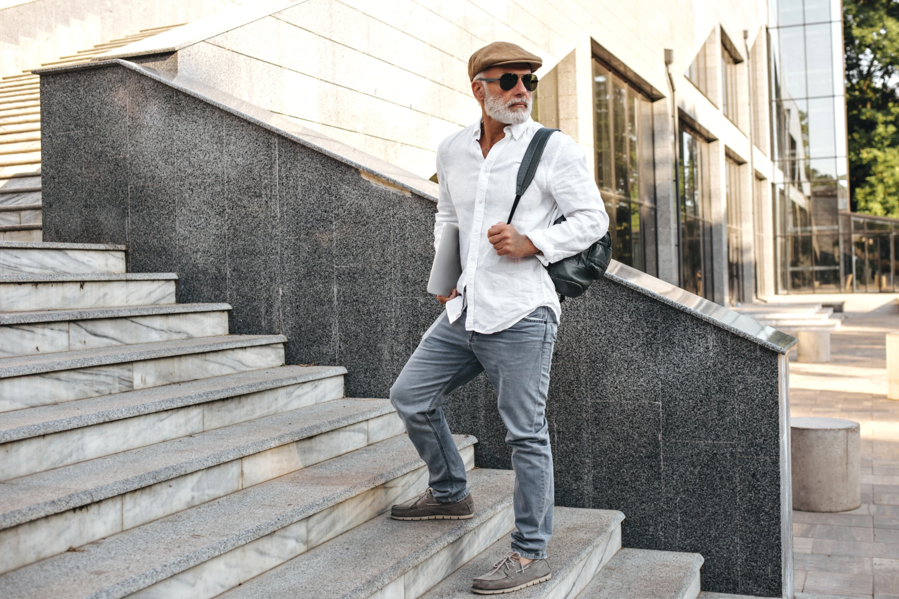 Man wearing a shirt, jeans, a cap, goggles, and a bag, walking up some steps in front of a building