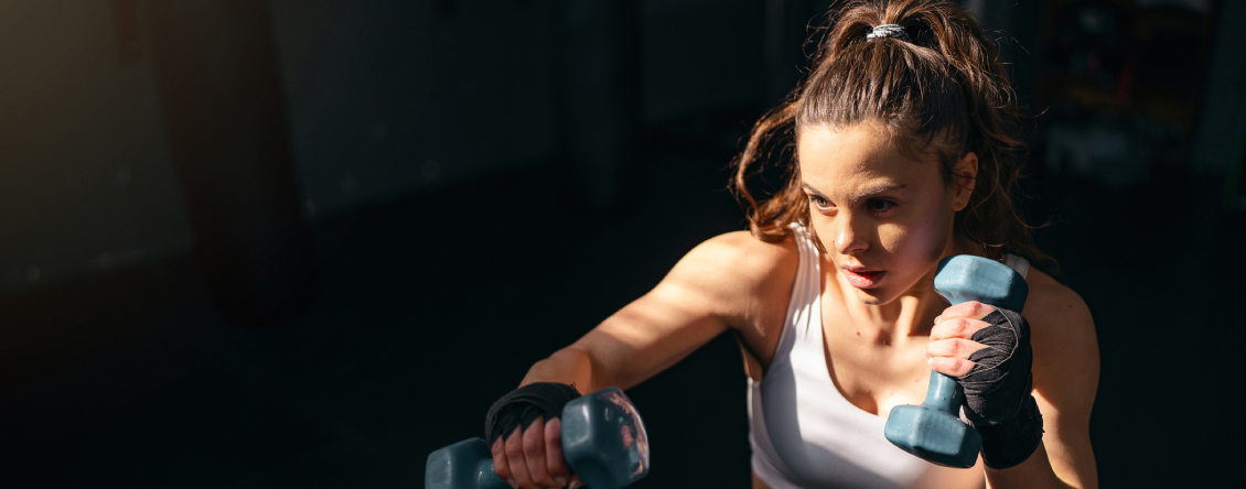 Woman in a white tank top and black shorts lifting two dumbbells in a dark gym