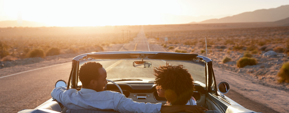 Man and woman in a convertible car driving down a desert road, surrounded by plants on either side